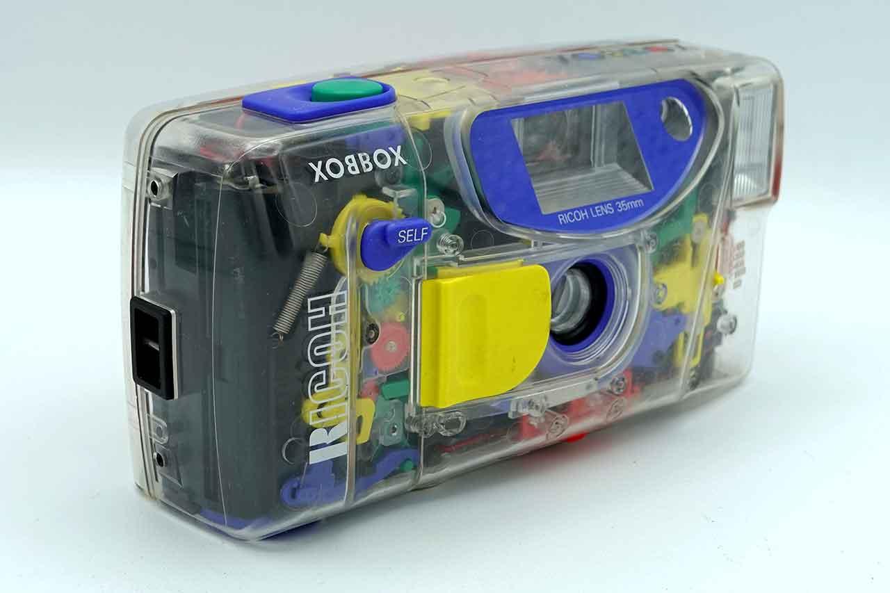 A transparent Ricoh 35mm film camera with its colourful internal gears, springs and film transport visible.